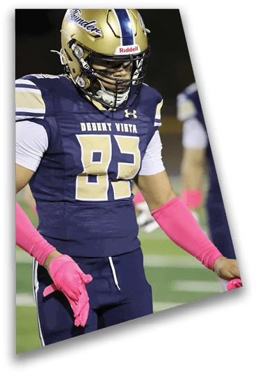 A person wearing a gold football helmet, navy blue and white uniform with the number 83, and bright pink gloves is standing on a football field. Description generated by AI