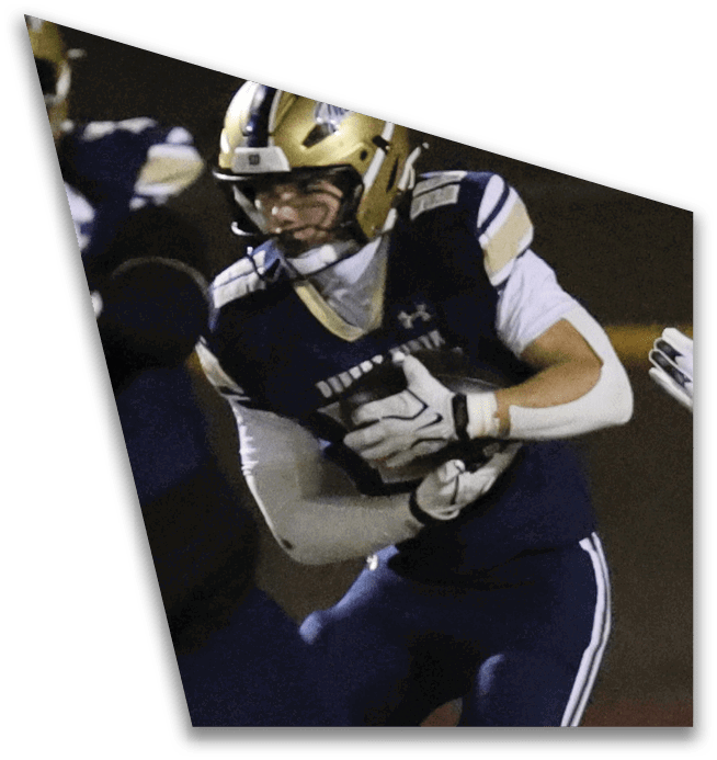 People in football uniforms are engaged in a play on a field at night, with one person holding the ball and others reaching out. Description generated by AI