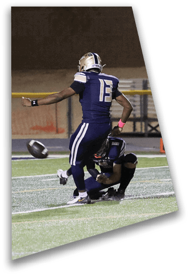 A person in a dark sports uniform is kicking a football held by another person crouching on a field at night. Description generated by AI