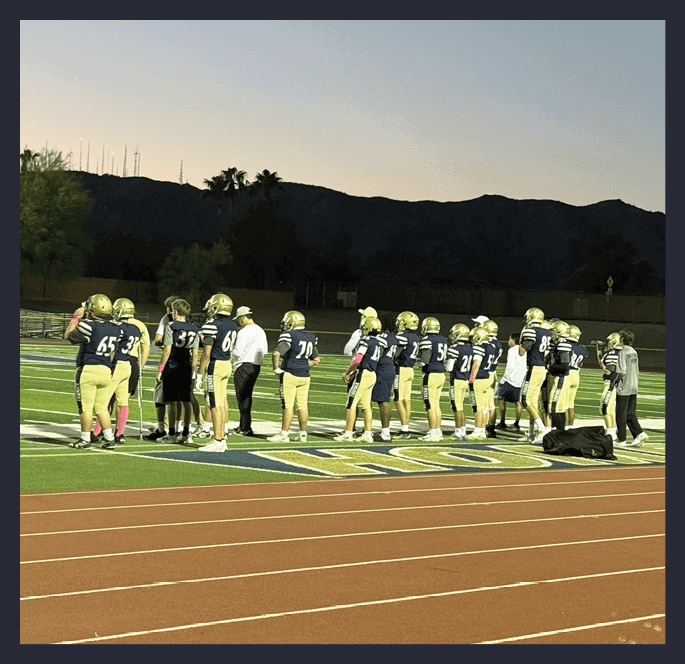 People in football uniforms and coaches stand on a field at dusk with stadium lights on and mountains in the background. Description generated by AI