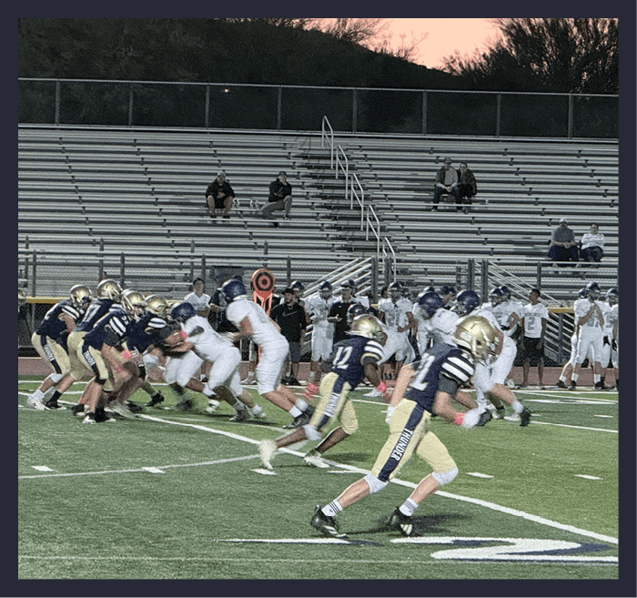 A group of people wearing football uniforms are engaged in a game on a field with empty bleachers and a sunset sky in the background. Description generated by AI
