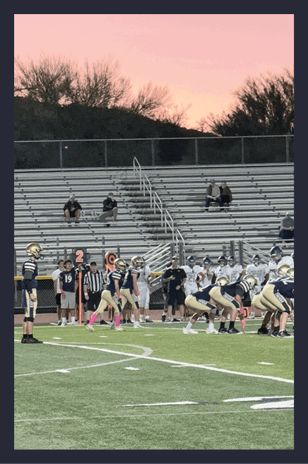 A group of people in sports uniforms are gathered on the sidelines of a football field with empty bleachers and a sunset sky in the background. Description generated by AI