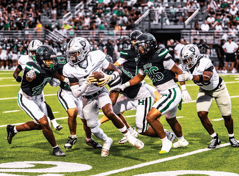 Four people wearing black football uniforms with “VISTA" on the front are standing together in a stadium with a crowd in the background, two of them holding helmets raised above their heads. Description generated by AI