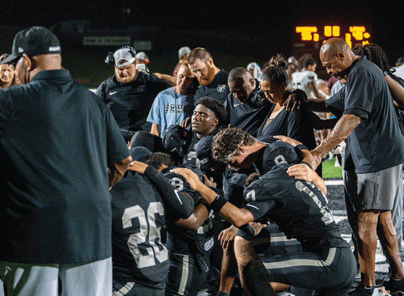 Four people wearing black football uniforms with “VISTA" on the front are standing together in a stadium with a crowd in the background, two of them holding helmets raised above their heads. Description generated by AI