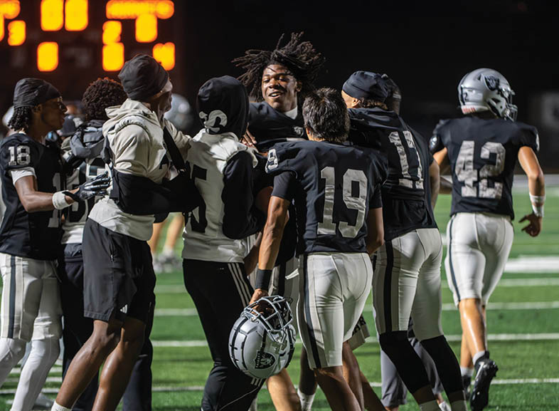Four people wearing black football uniforms with “VISTA" on the front are standing together in a stadium with a crowd in the background, two of them holding helmets raised above their heads. Description generated by AI