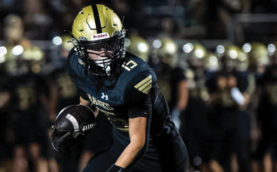 A person wearing a gold helmet and dark sports uniform is running with a football on a field with blurred players in the background. Description generated by AI
