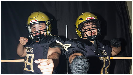 Three people wearing football helmets and black shirts with “HAWKS" printed on them are holding and eating pizza from an open pizza box. Description generated by AI