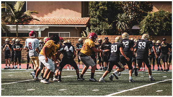 People wearing helmets and sports uniforms are playing football on a field with houses and trees in the background. Description generated by AI