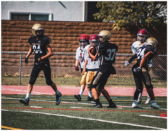 Five people wearing football helmets and uniforms are standing on a football field with a brick wall and trees in the background. Description generated by AI