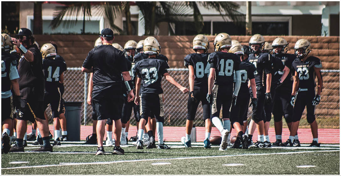 A group of people wearing black football uniforms and gold helmets are gathered on a field near a chain-link fence with a brick wall and palm trees in the background. Description generated by AI