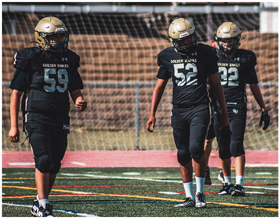 Three people wearing black football uniforms and gold helmets walk on a sports field with a goal net in the background. Description generated by AI