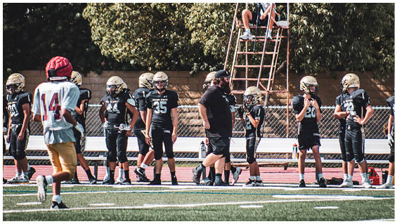 People in football uniforms and a coach stand on a field near a track with a person sitting on a raised platform in the background. Description generated by AI