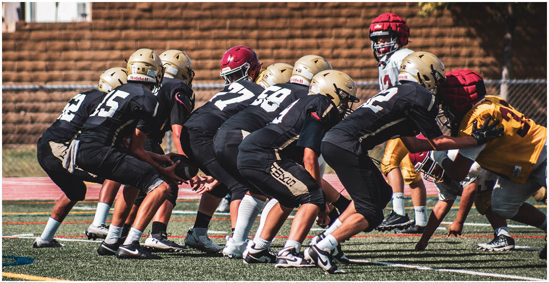 A group of people wearing black and gold sports uniforms are lined up on a football field facing opponents in red and yellow uniforms during a game. Description generated by AI