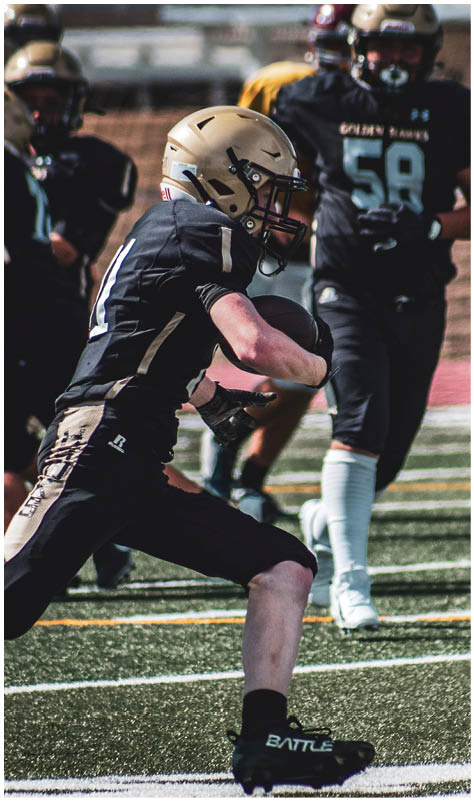Two people wearing black football uniforms and gold helmets are on a football field, with one person crouching and the other standing behind. Description generated by AI