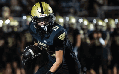A person wearing a gold helmet and dark sports uniform is running with a football on a field with blurred players in the background. Description generated by AI