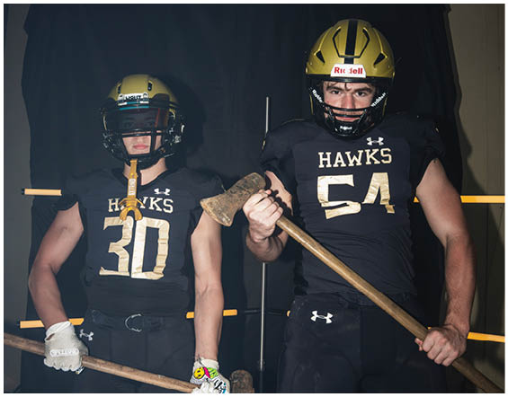 Two people wearing black football uniforms and gold helmets stand side by side, with one holding an axe. Description generated by AI