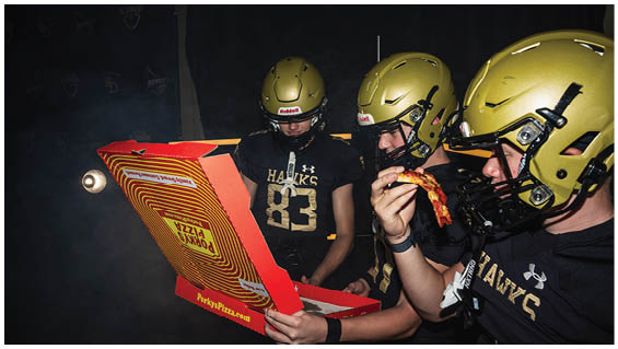 Three people wearing football helmets and black shirts with “HAWKS" printed on them are holding and eating pizza from an open pizza box. Description generated by AI