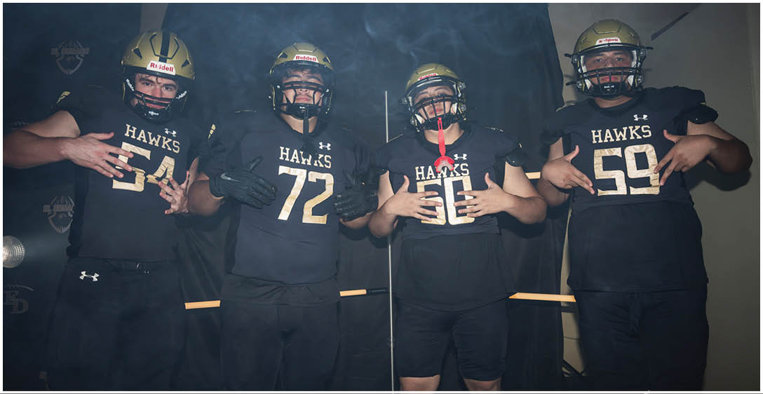 Four people wearing black football uniforms with gold helmets and the word Hawks on their jerseys pose together in a smoky environment. Description generated by AI
