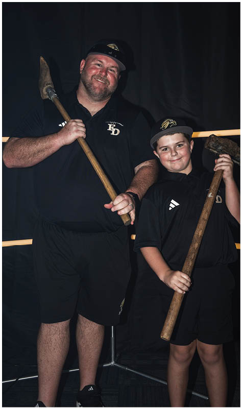 Two people wearing matching black shirts and hats are smiling and holding wooden tools with metal ends against a dark background. Description generated by AI