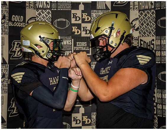 Two people in black football uniforms and helmets face each other with their fists pressed together in front of a backdrop featuring football-related text and logos. Description generated by AI