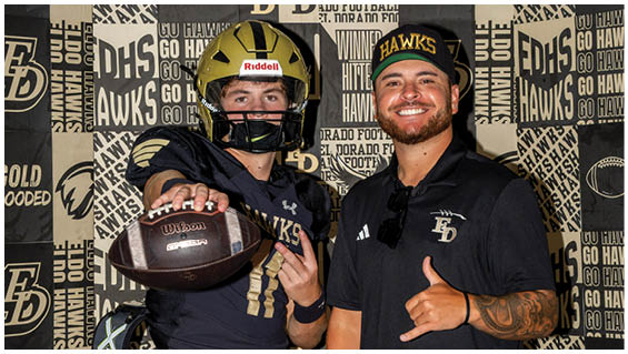 Two people wearing sports attire pose in front of a backdrop with football-themed text and logos, with one person holding a football and the other giving a thumbs-up. Description generated by AI