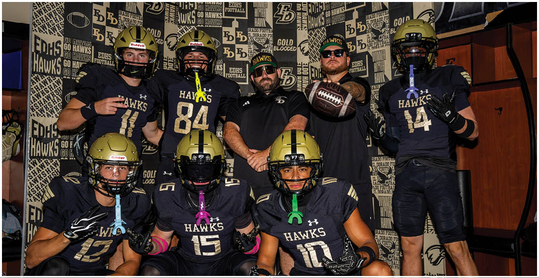 A group of adults wearing black football uniforms with gold helmets pose together indoors against a backdrop with team logos and slogans. Description generated by AI