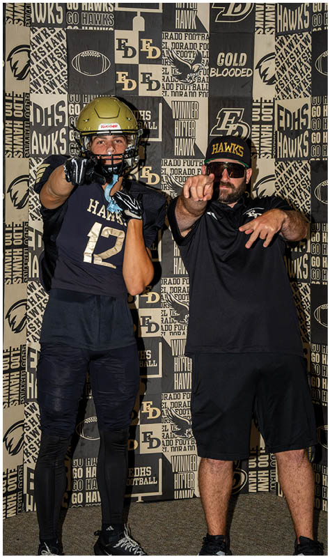 A person wearing a football helmet and uniform stands next to a person in sunglasses and a cap pointing forward against a backdrop with football-themed text and logos. Description generated by AI
