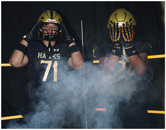 Two people wearing black football uniforms and gold helmets pose with one holding the helmet and the other covering their face with gloved hands amid smoke. Description generated by AI