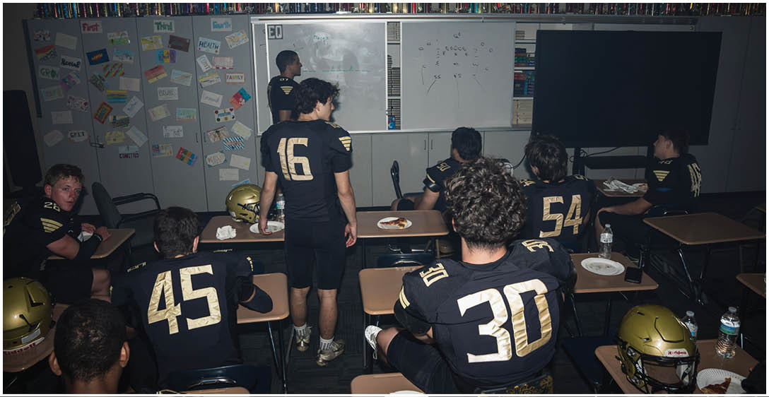 People wearing football uniforms are seated and standing in a classroom while looking at a person writing on a whiteboard. Description generated by AI