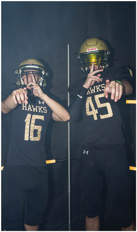 Two people wearing black football uniforms with gold helmets and the word “HAWKS" on their jerseys pose with hand gestures against a dark background. Description generated by AI