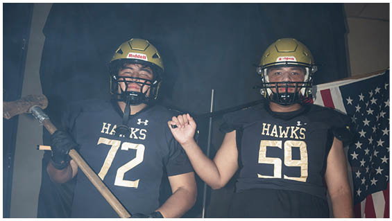 Two people wearing football helmets and black jerseys with the numbers 72 and 59 stand side by side, one holding a sledgehammer and the other raising a fist, with an American flag partially visible in the background. Description generated by AI