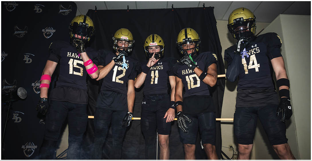 Five people wearing black football uniforms and gold helmets pose together indoors against a dark backdrop. Description generated by AI