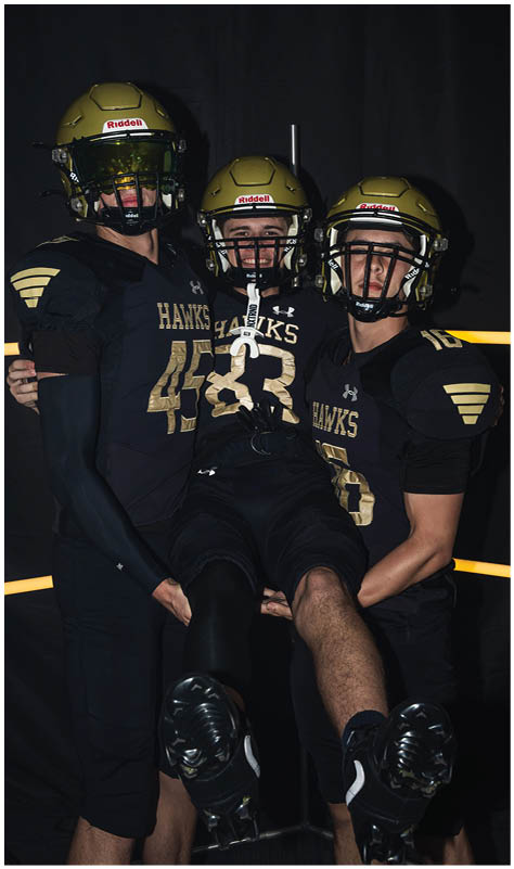 Three people wearing black and gold football uniforms and helmets are posing together with two standing and holding the third person horizontally between them against a dark background. Description generated by AI