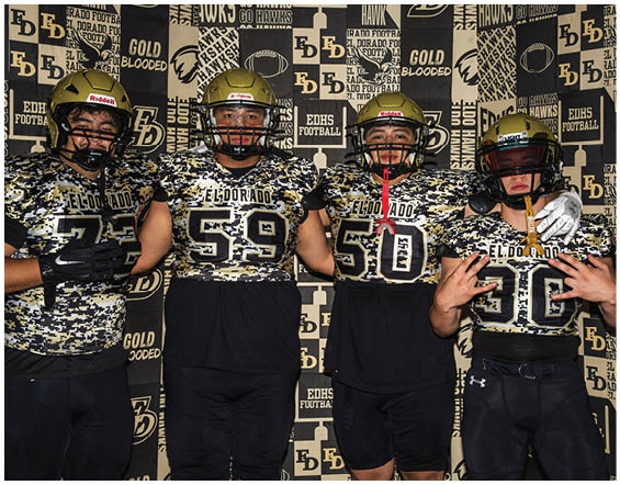 Four people wearing black athletic shorts and matching black and gold sports jerseys stand side by side against a backdrop with repeated text and logos. Description generated by AI