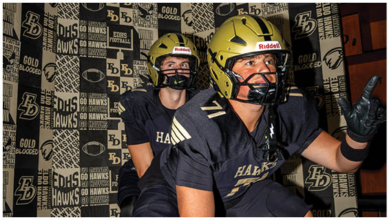 A wall covered with patterned banners featuring the text “DHS," "EDHS," "GO HAWKS," "EXEDS FOOTBALL," and "GOLD BLOODED" along with a stylized hawk logo. Description generated by AI