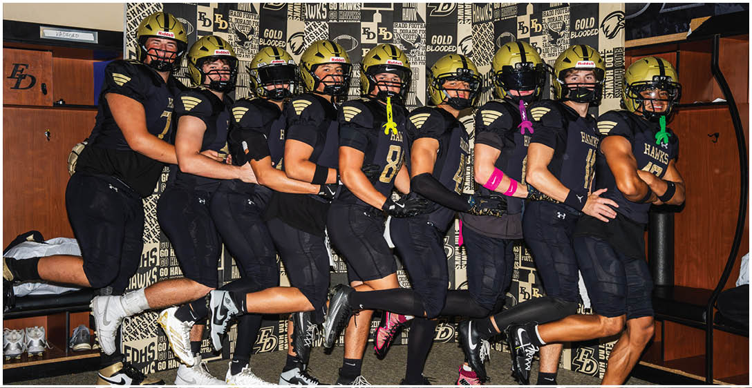 A group of people wearing black football uniforms and gold helmets stand in a line with one leg raised, posing energetically in a locker room. Description generated by AI