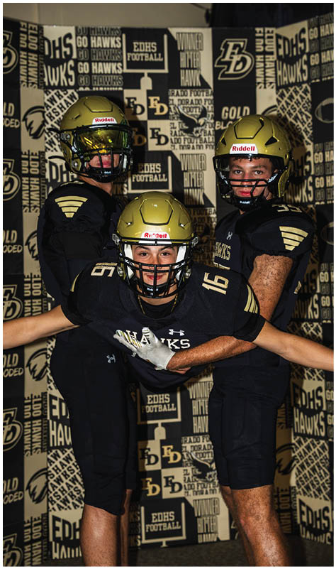 Three people in black football uniforms and gold helmets pose together in front of a backdrop with football-related text and logos. Description generated by AI