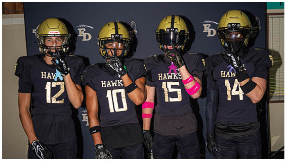 Four people wearing yellow football helmets stand in front of a black backdrop with repeated logos featuring a hawk, a shield, and the letters FD. Description generated by AI