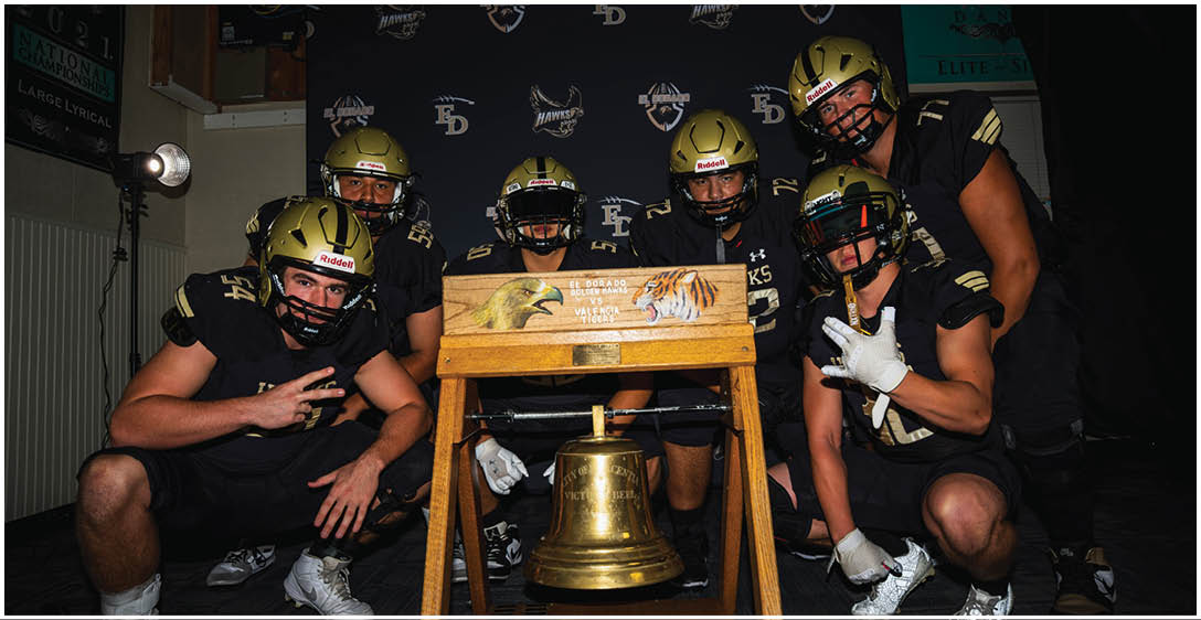 Five people in black sports uniforms and helmets pose around a wooden stand with a large brass bell in front of a backdrop with logos. Description generated by AI