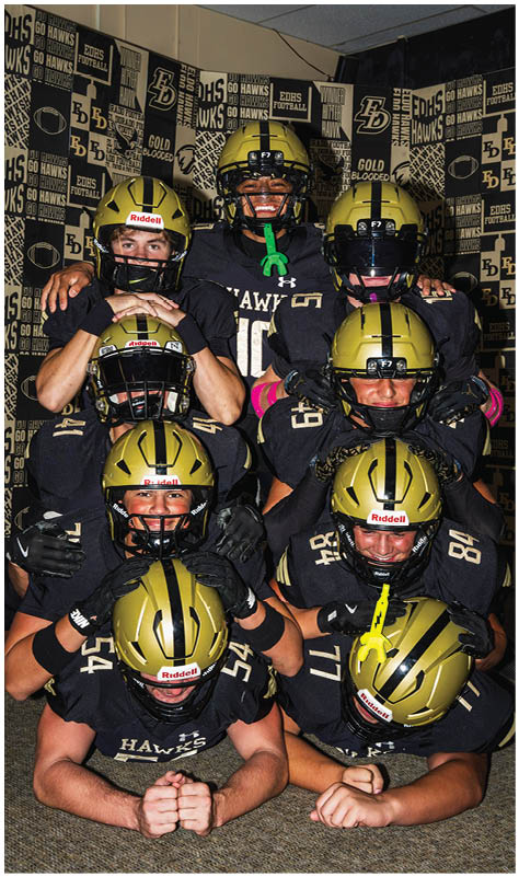 A group of people wearing black and gold football uniforms and helmets are stacked in a pyramid formation inside a room decorated with sports banners and posters. Description generated by AI