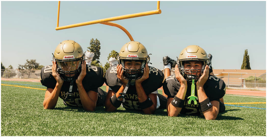 Three people wearing football helmets and black jerseys lie on the grass field under a goalpost, each holding their helmet with both hands. Description generated by AI