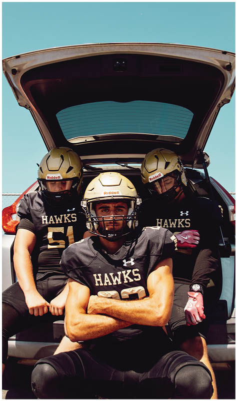 Three people wearing football helmets and black Hawks jerseys are sitting in the open trunk of a vehicle under a clear blue sky. Description generated by AI