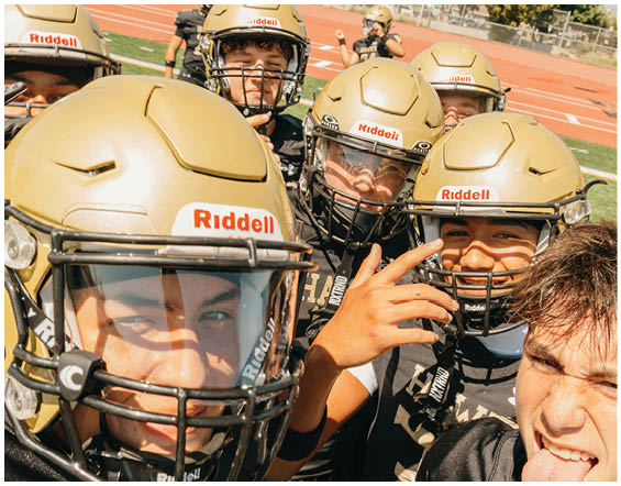 A group of people wearing gold football helmets and black jerseys are gathered closely together on a sports field, smiling and posing for a selfie. Description generated by AI