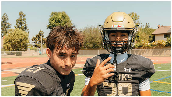 Two people wearing black football uniforms with numbers 27 and 38 are standing on a sports field. Description generated by AI