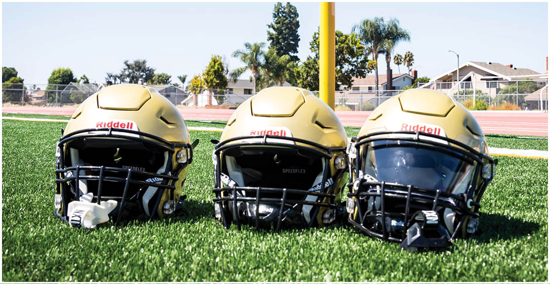 Three gold football helmets with black faceguards are placed on green turf with a track and houses visible in the background. Description generated by AI