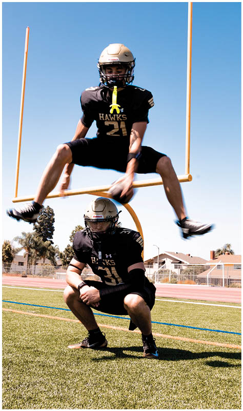 Two people in football uniforms and helmets are on a field with one person crouching and the other jumping over them through a goalpost. Description generated by AI