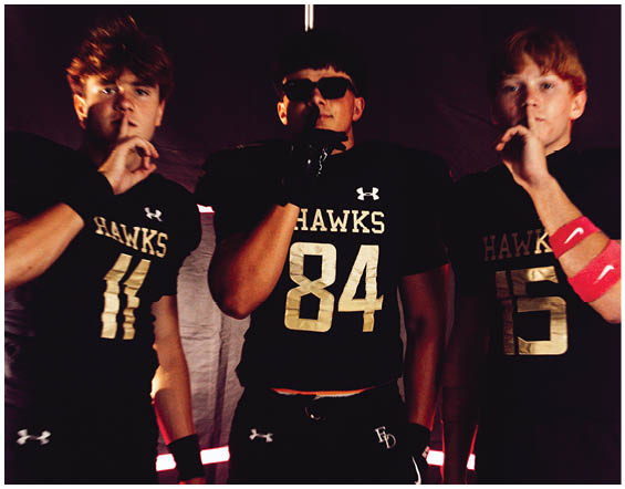 Three people wearing black football uniforms with “HAWKS" on the front pose with one finger held to their lips against a dark background. Description generated by AI