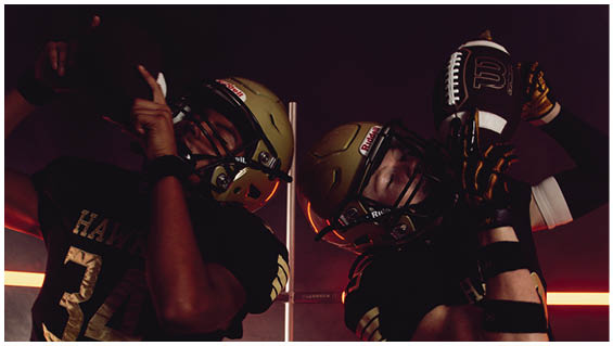 Two people wearing football helmets and black jerseys with gold numbers are posed in a dimly lit setting with red horizontal light accents. Description generated by AI