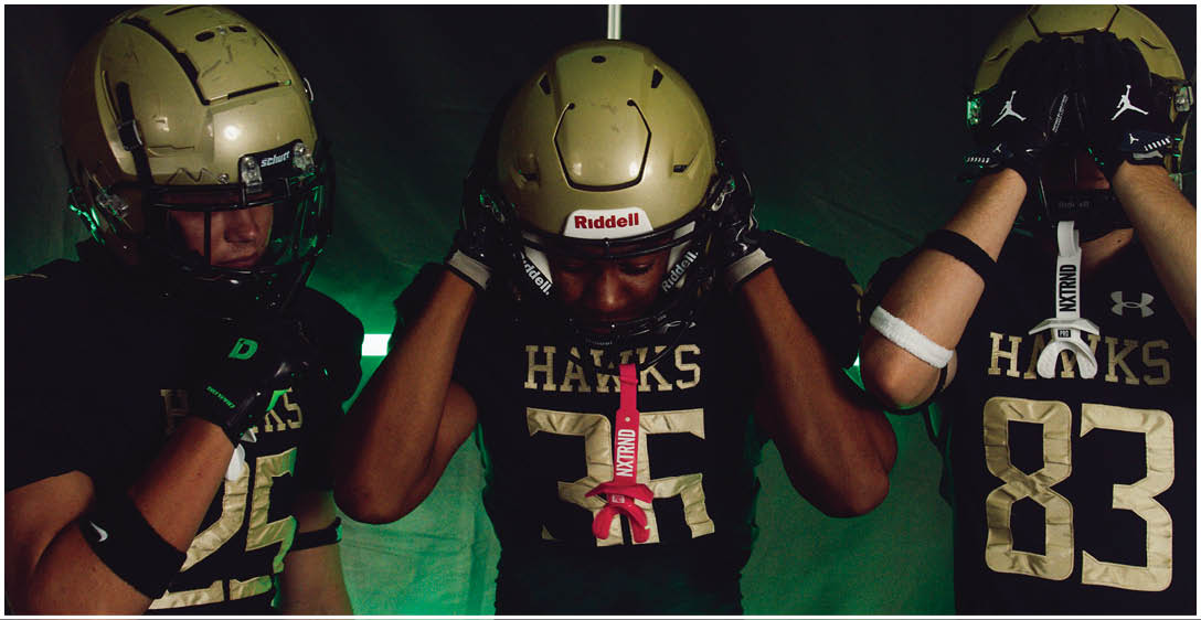 Three people wearing football helmets and black jerseys with the word “HAWKS" and numbers stand closely together against a dark green background. Description generated by AI