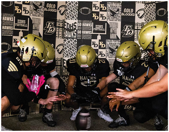 Four people wearing gold helmets and black football uniforms crouch around a football placed on the ground in front of a backdrop covered with sports-themed text and logos. Description generated by AI